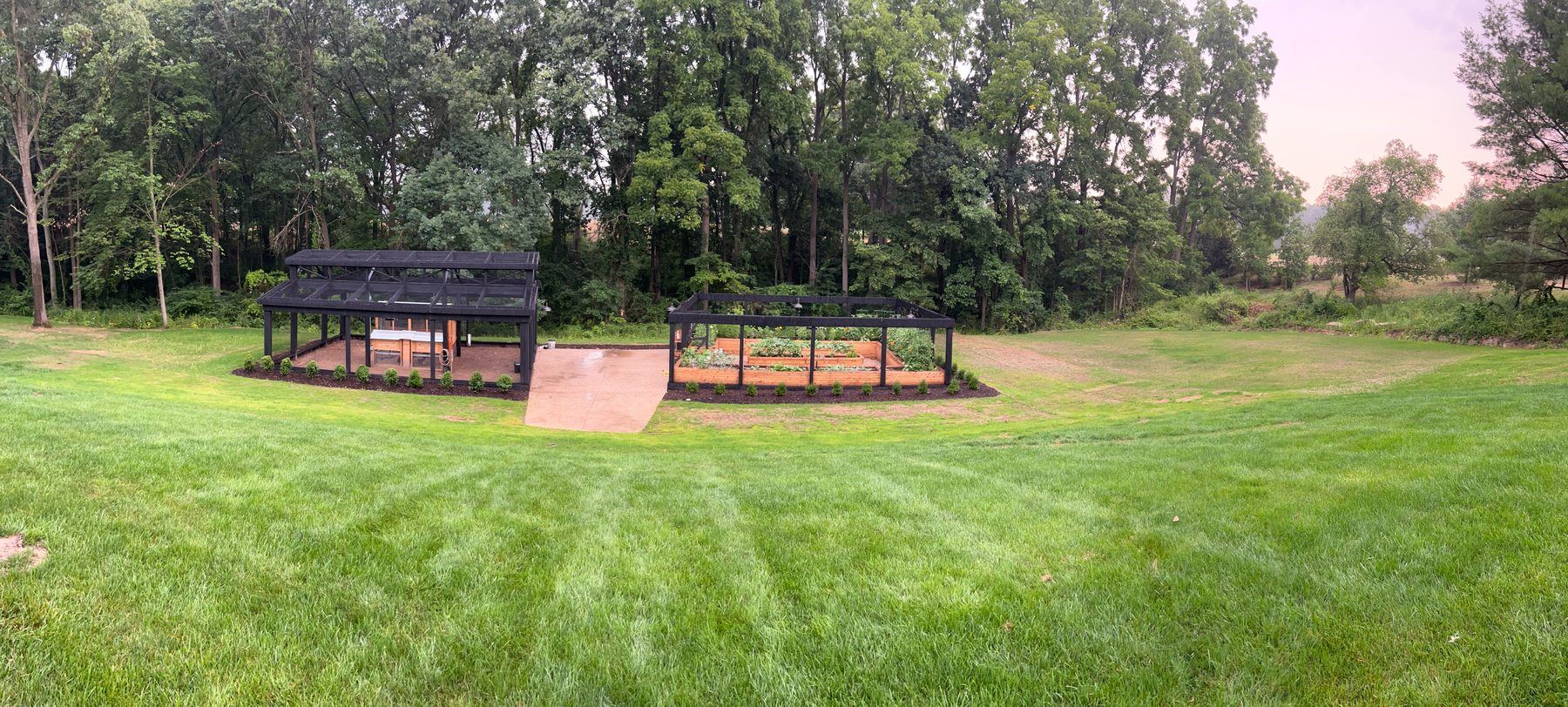 Panoramic view of black timber coop and enclosed garden compound on a green lawn against a treeline